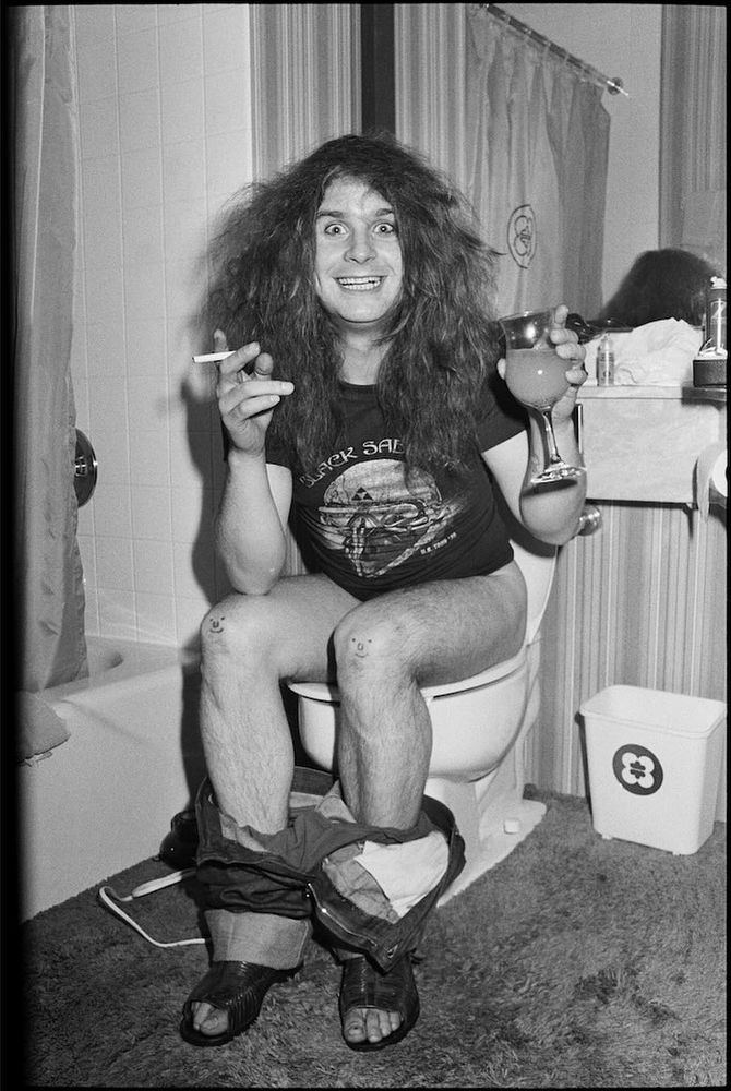 Ozzy Osbourne in a Black Sabbath T-shirt, with his pants down, sitting on a toilet with a drink in one hand and a cigarette in the other. He's got wide eyes and a manic smile.