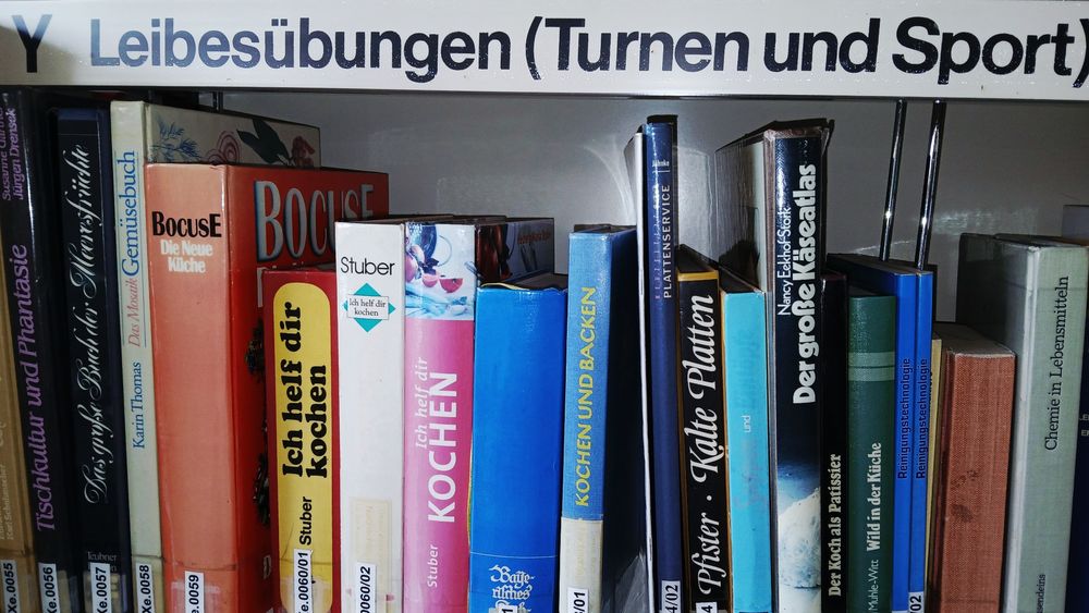 Das Bild zeigt ein Bibliotheksregal mit der Beschriftung „Y Leibesübungen (Turnen und Sport)”. Trotz dieser Kategorie befinden sich auf dem Regalbrett überwiegend Kochbücher und Bücher rund um das Thema Essen und Küche, zum Beispiel Titel wie „Ich helf dir kochen“, „Kochen und Backen“ oder „Der große Käseatlas“. Die Bücher stehen dicht aneinander gereiht, einige mit bunten, andere mit schlichten Einbänden.