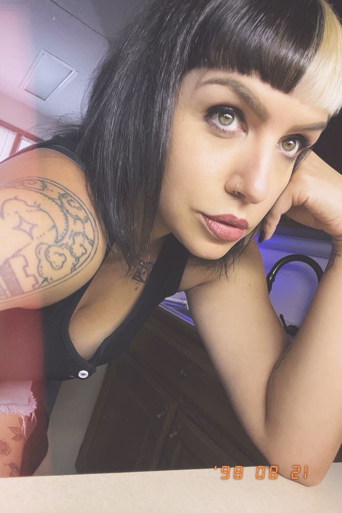 A selfie of me looking up towards the light. I’m in a black tank top. You can see a tattoo on my shoulder. I have dark hair with bleached split-dyed bangs