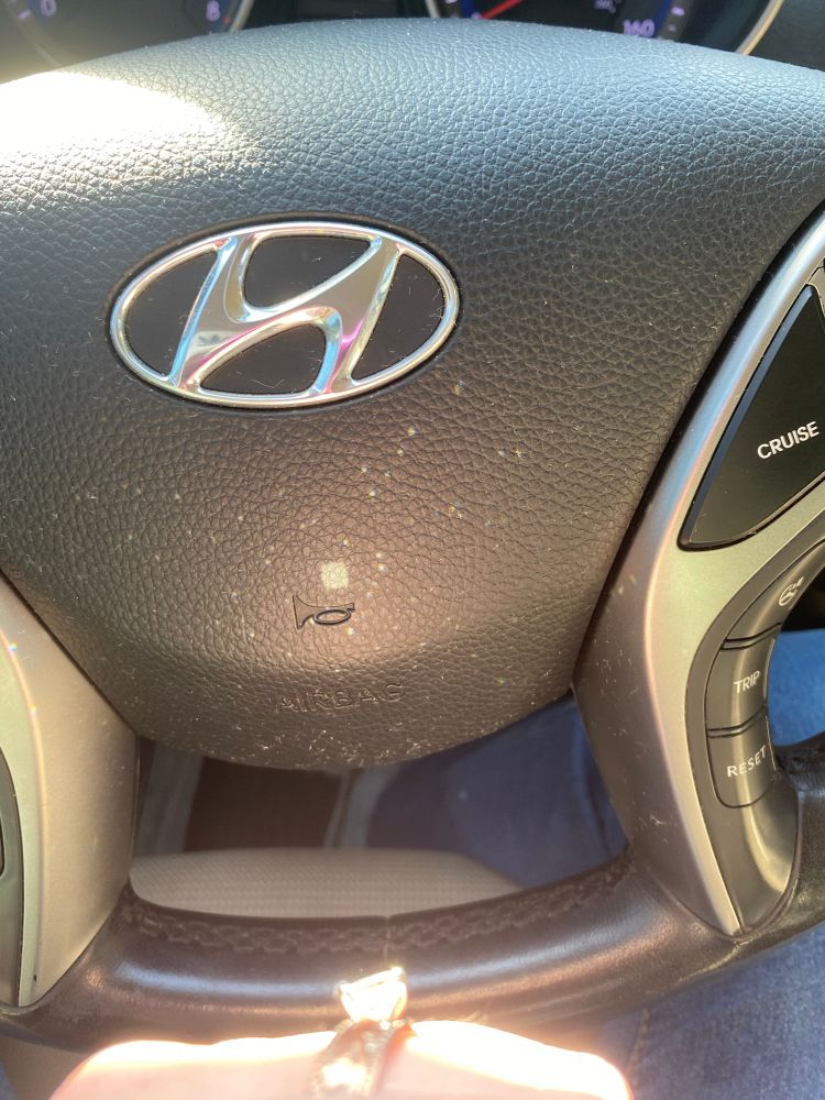 A hyundai steering wheel, but at the bottom is my engagement ring lit up in the sunlight, and reflecting lots of little sparkles and one perfect square onto the black leather 