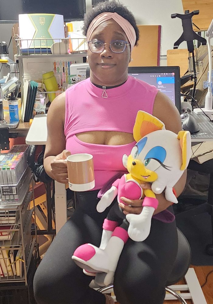 Photograph of myself holding a Rouge the Bat plush and brown Pantone coffee mug while sitting at my workstation. I have a worried smiling expression on my face.
