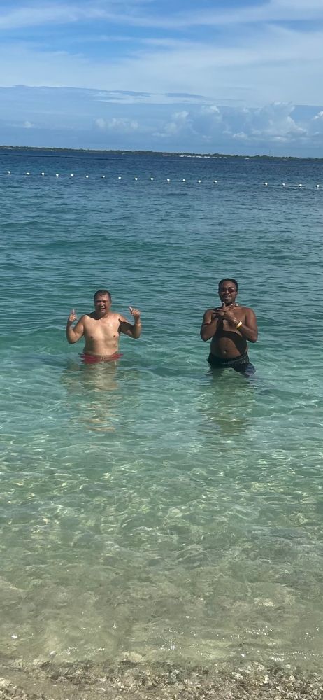 The two men stand facing each other in the shallow water near the sandy shore. The sky is clear, and boats can be seen in the distance.