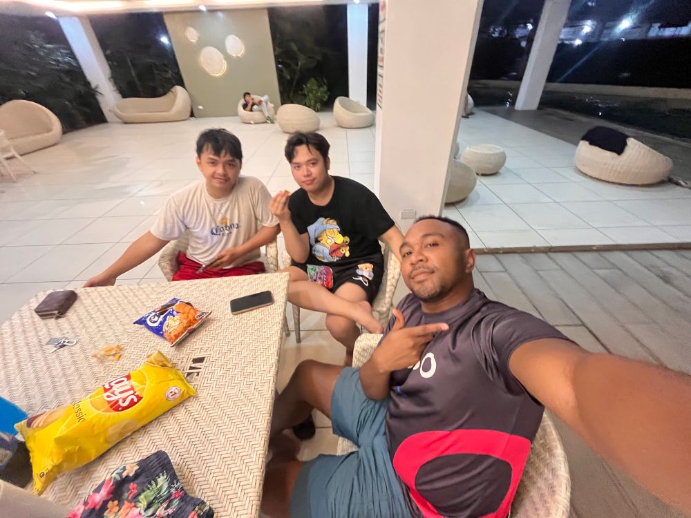 Three men sitting at a table in an outdoor lounge area at night, taking a selfie. The man on the right, wearing a black and red sports shirt, holds the camera and makes a hand gesture. The man in the middle wears a black graphic T-shirt and holds a snack. The man on the left, in a white T-shirt and red shorts, leans on the table with a relaxed smile. There are bags of chips and snacks on the table, and the background features white lounge chairs and warm lighting.