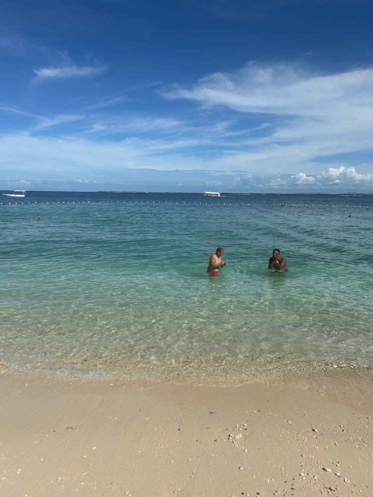 Two men standing in clear, waist-deep turquoise water at a beach. One wears red swim trunks, and the other wears black trunks, both making hand gestures.