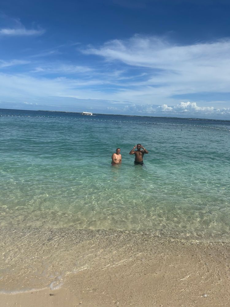 A wider shot of the same two men in shallow turquoise water. One has red swim trunks, the other black, with a white boat visible on the horizon.