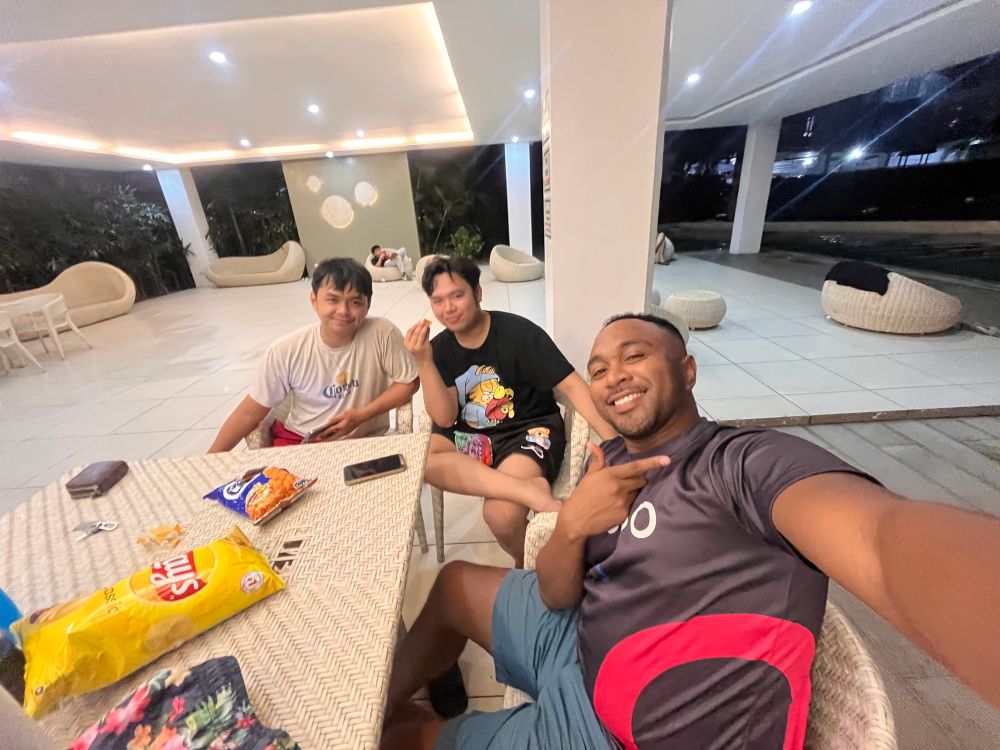 Three men sitting at a table in an outdoor lounge area at night, taking a selfie. The man on the right, wearing a black and red sports shirt, holds the camera and makes a hand gesture. The man in the middle wears a black graphic T-shirt and holds a snack. The man on the left, in a white T-shirt and red shorts, leans on the table with a relaxed smile. There are bags of chips and snacks on the table, and the background features white lounge chairs and warm lighting.