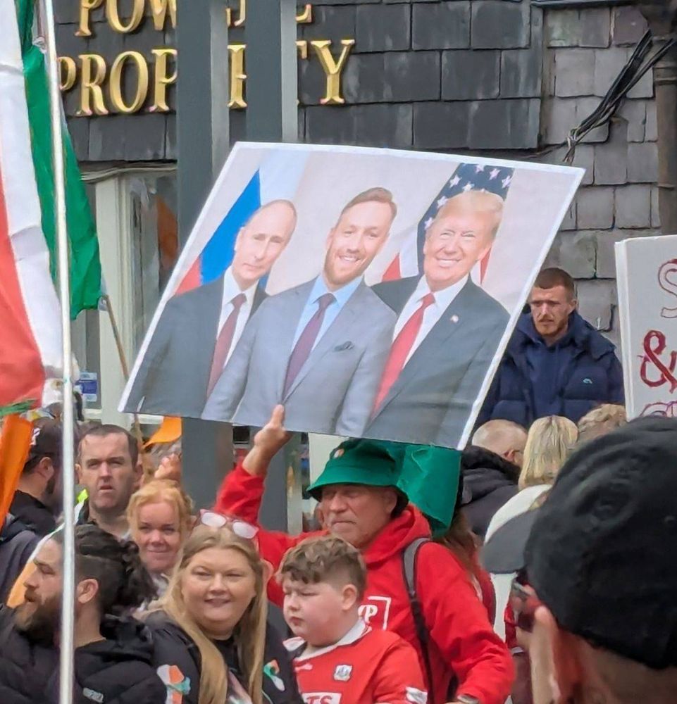 Far right protester holding large sign with Vladmimir Putin, Conor McGregor, and Donald Trump standing together and smiling
