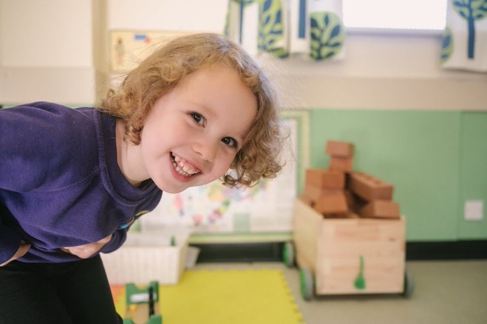 One of the Naíscoil kids leaning and looking into the camera lens. The background is the pre-school classroom. 