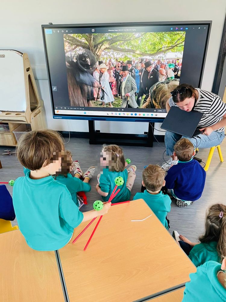 Photo of our Naíscoil leader Cíara showing the children some pictures of her trip to the Buckingham Palace garden party. The children and another teacher are sitting on little chairs and tables.