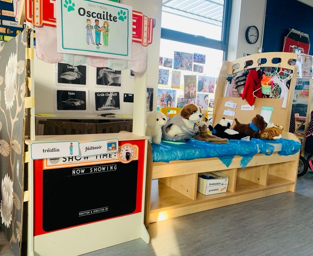Ár gclinic tréidlia! Our vet clinic. Photo of stuffed animals set up as if they are at the vets. There are boxes for bandages and plasters labelled in Irish. A sign says Oscailte - Open. 