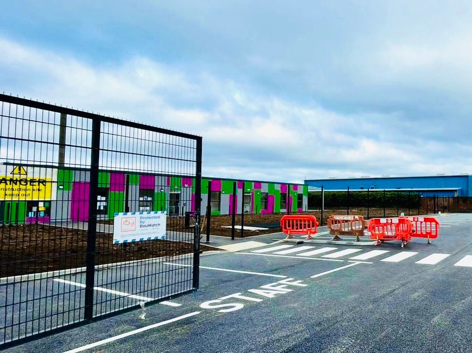 Photo of Scoil na Seolta’s new building. It’s single storey, has purple and green panels and big windows. You can also see the school gate and staff parking spaces. 