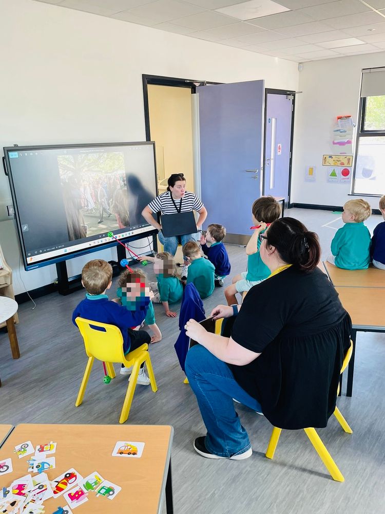 Photo of our Naíscoil leader Cíara showing the children some pictures of her trip to the Buckingham Palace garden party. The children and another teacher are sitting on little chairs and tables.
