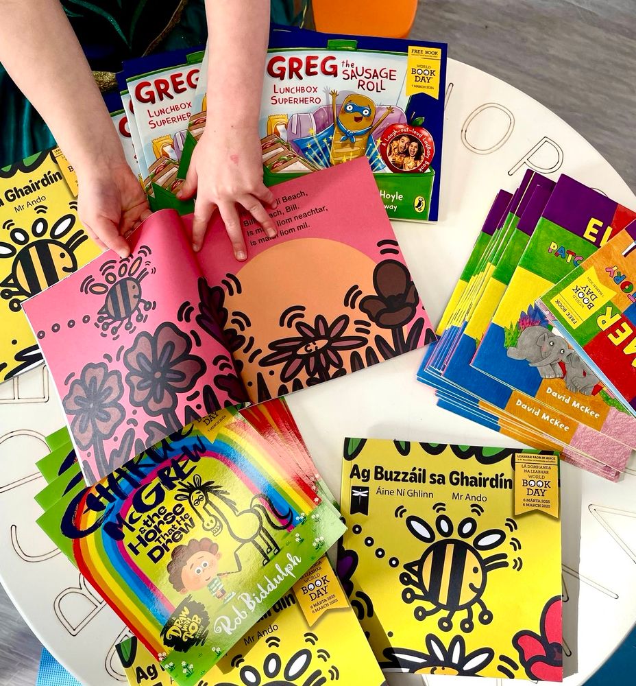One of the kids’ hands opening a book, with a table full of Irish language children’s books 