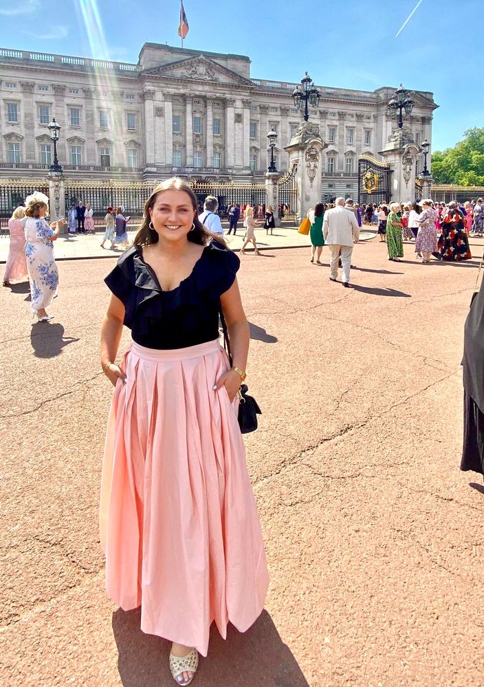 Cíara wearing a long pink skirt and black top in the sunshine outside Buckingham Palace. 