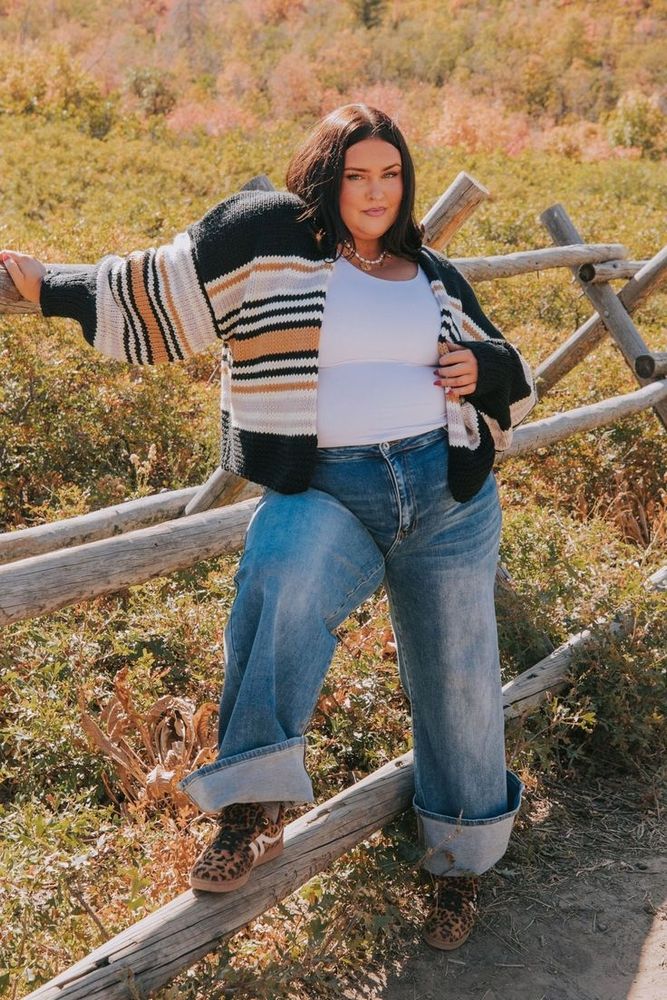 A model making a pose on a wooden fence in a field. They are wearing a white tank top with a gold necklace I think. A striped cardigan sweater thing with black, white, tan and orange. And a blue jeans a cuff I think it's called. Also some fancy shoes I think. I don't. I'm not good with fashion so I don't know. I try my best. Have a good day. You're beautiful and I love you. 