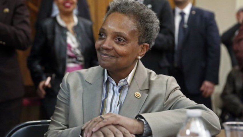 Lori Lightfoot in one of her grey overly large suits, with her hands folded and a pleased and bemused expression her somehow always memeable face 