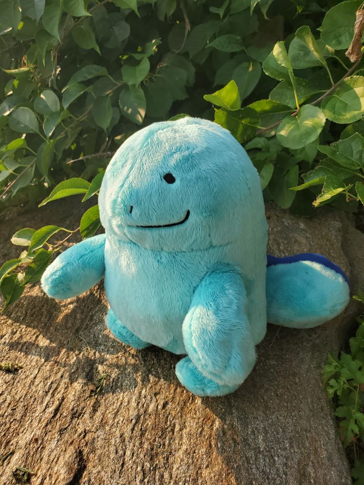 Handmade plush of the pokemon Quagsire sitting on a rock in front of green bushes.
