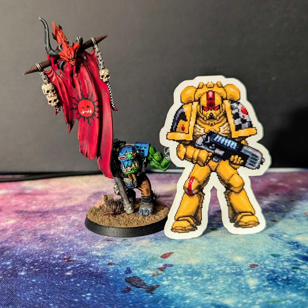 A pixel art space marine sticker propped up by an ork miniature.