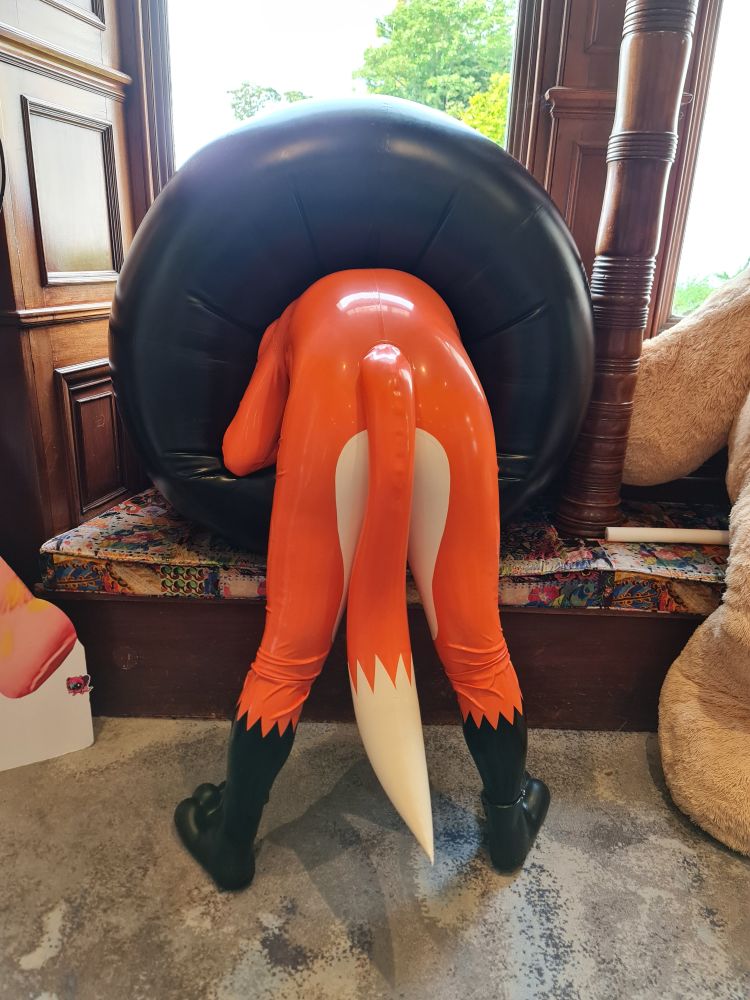 A photograph of a person wearing a rubber orange, white and black costume. They are facing away from the camera and they are leaning through a large black inflatable ring.