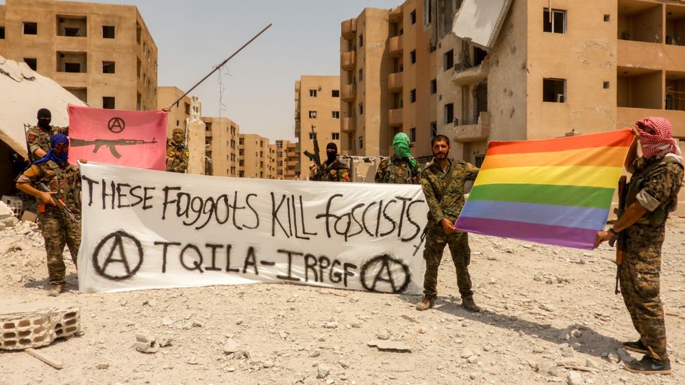 TQILA (International Revolutionary People's Guerrilla Forces) forces in Raqqa, Syria, posing with a rainbow flag and a plane saying "These faggots kill fascists" with anarchy symbols and the name of the unit

