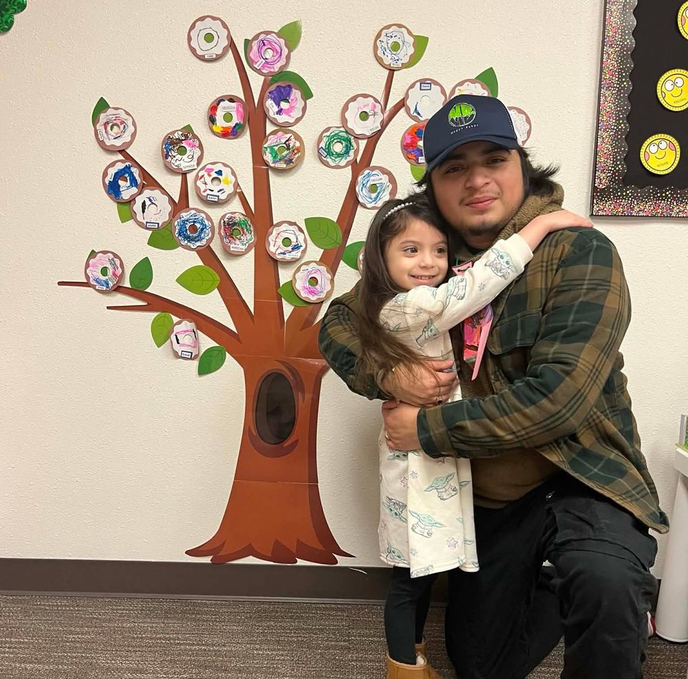 Past photo of a smiling Wilmer kneeling to hug his smiling daughter in front of school kid art. 
