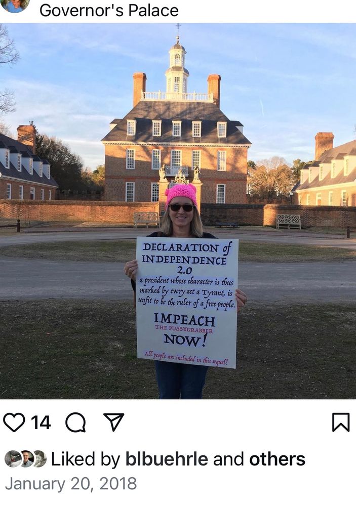 Woman in pink knit hat standing in front of colonial governor’s palace holding sign. 