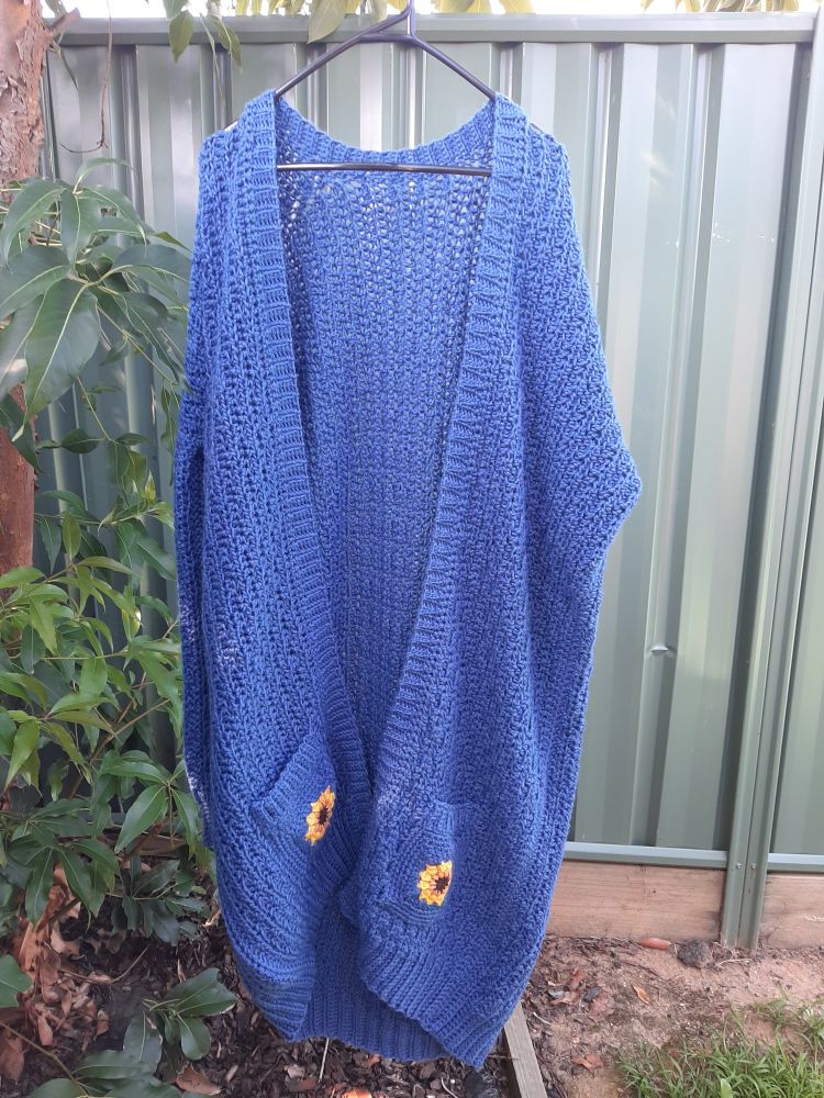 a long, blue crocheted cardigan with pockets at the bottom with yellow sunflowers on them. its hanging on a coat hanger in front of a fence. 