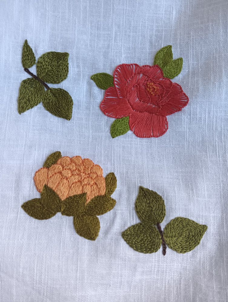 Tela de lino blanca en la que están bordadas dos flores y dos ramitas de hojas. Una flor está bordada en punto long and short, color amarillo bordeada con un naranja que le complementa. La rodean siete hojitas verdes en punto atrás completo. La otra flor está bordada en rojo, punto festón y sus hojitas son de otro verde diferente, también en atrás completo. Las dos ramitas de hojas están bordadas en punto atrás completo el tallo, que es marrón y las hojas en punto cadeneta, son de otro tono verde, diferente al de las hojas de las flores. 