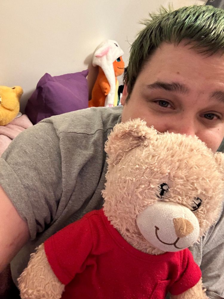 A white trans woman with short green hair, they are holding a light brown build a bear in front of their mouth, they are wearing a light grey t-shirt and the bear is wearing a red t-shirt.