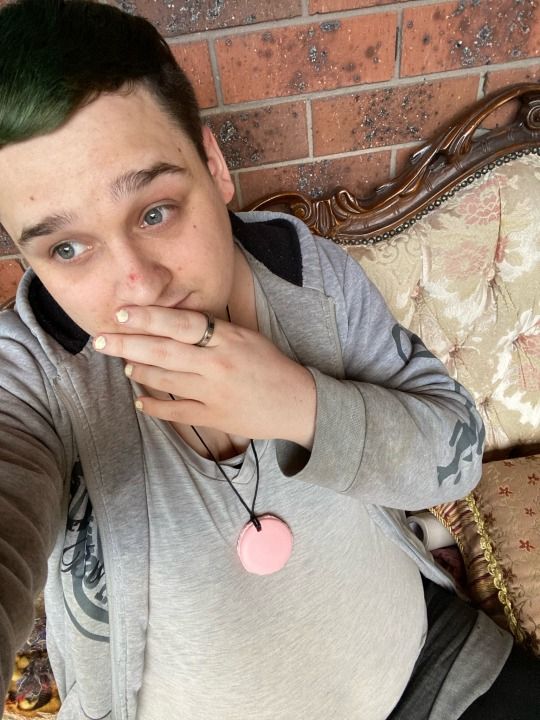 A trans person sitting on a couch in front of a brick wall, they have short green hair and are wearing yellow nail polish.