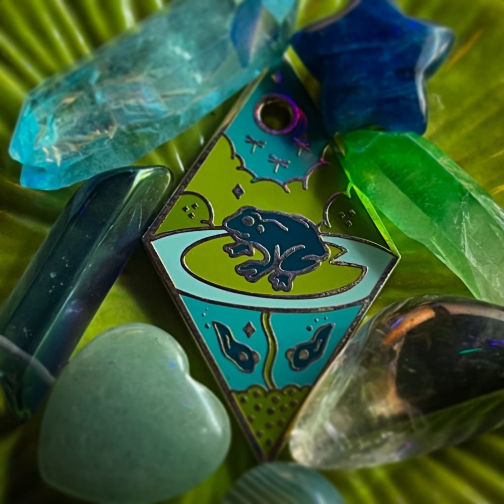“The Frog” Diamond shaped enamel charm with the image of a frog on a lily pad; under the water are two tadpoles and a mound of frog eggs; overhead in the sky are golden dragonflies - the tile is surrounded by semi-precious stones in blue, green, and teal hues. The lighting is cool and dark. The background is a ceramic lily pad incense burner.