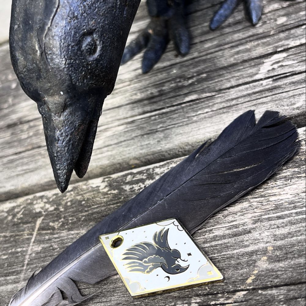 A cast iron crow looks down at a diamond shaped enamel charm that is inlaid with the image of a crow in flight. The charm is on a black pigeon feather. The whole image is backed by a plane of weathered wood.
