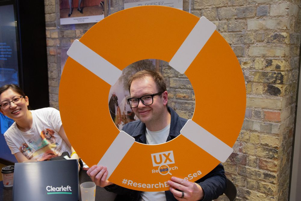 A man wearing glasses and a dark jacket smiles while holding up an orange lifebuoy prop branded with "UX Research by the Sea" and the hashtag #ResearchByTheSea. A woman in a white t-shirt sits behind him, also smiling. The background features a brick wall with framed historical posters.