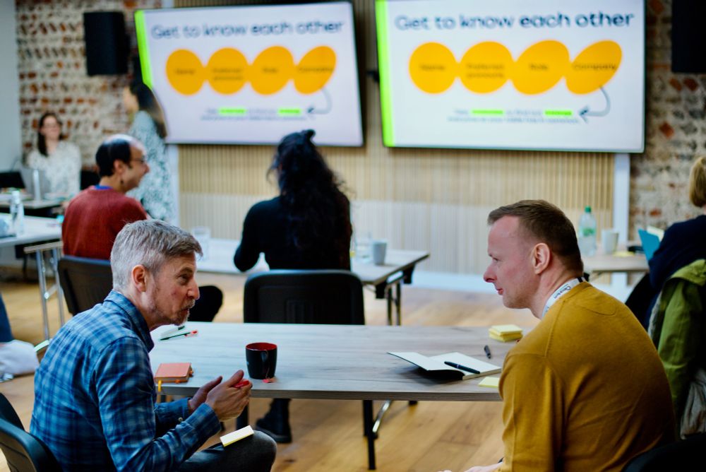 Two participants in a workshop are seated at a table, engaged in a one-to-one discussion. A large presentation screen in the background displays the words "Get to know each other" along with a diagram. Other attendees are visible in the background.