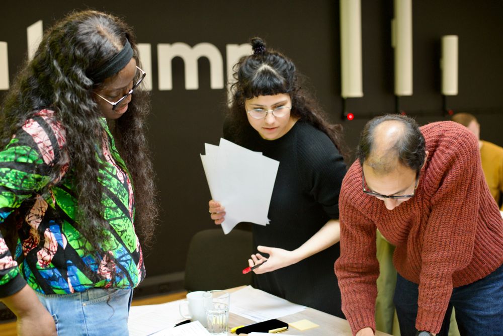 Three people engaged in an active discussion over a table with papers and sticky notes. One woman holds a few sheets of paper while explaining something to the others, who are leaning in attentively.
