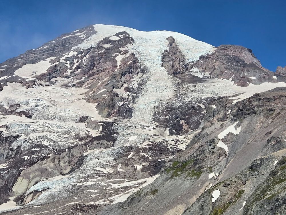 A rugged perspective of Mount Rainier’s upper face from the Skyline Trail, where jagged cliffs and dark volcanic rock meet sprawling glaciers and patches of lingering snow. The sheer scale of the icy slopes against the rocky terrain highlights both the mountain’s power and its raw beauty.
