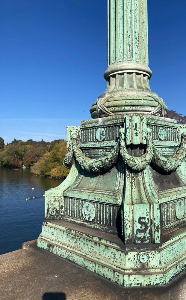 The number 5 appears on the base of an early 19th-century lamppost on top of a bridge. Five swans can be seen on the dark-blue river, flanked by green trees with the odd speck of autumn reds and golds. 