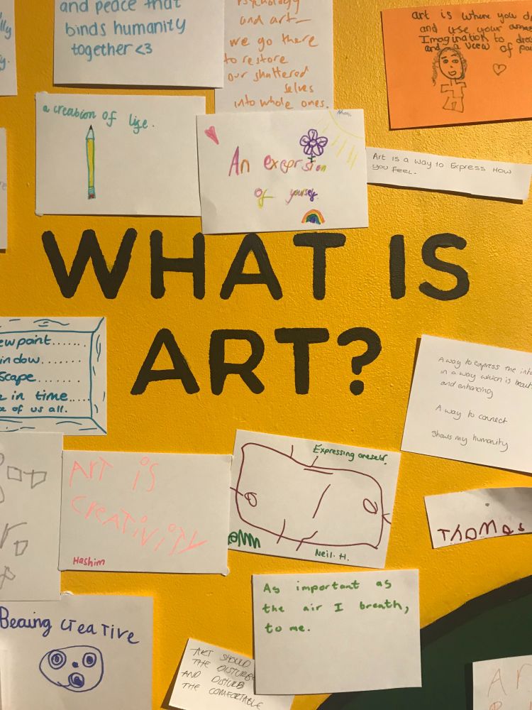 An yellow wall with the text, "What is art" written in black. Pinned to the yellow wall are various audience responses written on white paper. The pieces of paper say 'As important as the air I breathe to me,' 'Expressing oneself,' 'a creation of life.'  
