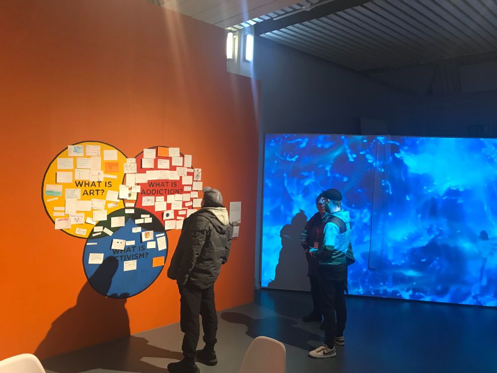 Two people are standing in front of an interactive wall display with three overlapping circles labelled "What is Art?", "What is Addiction?", and "What is Activism?". The circles are filled with colourful handwritten notes. To the right, a bright blue video projection illuminates the space.