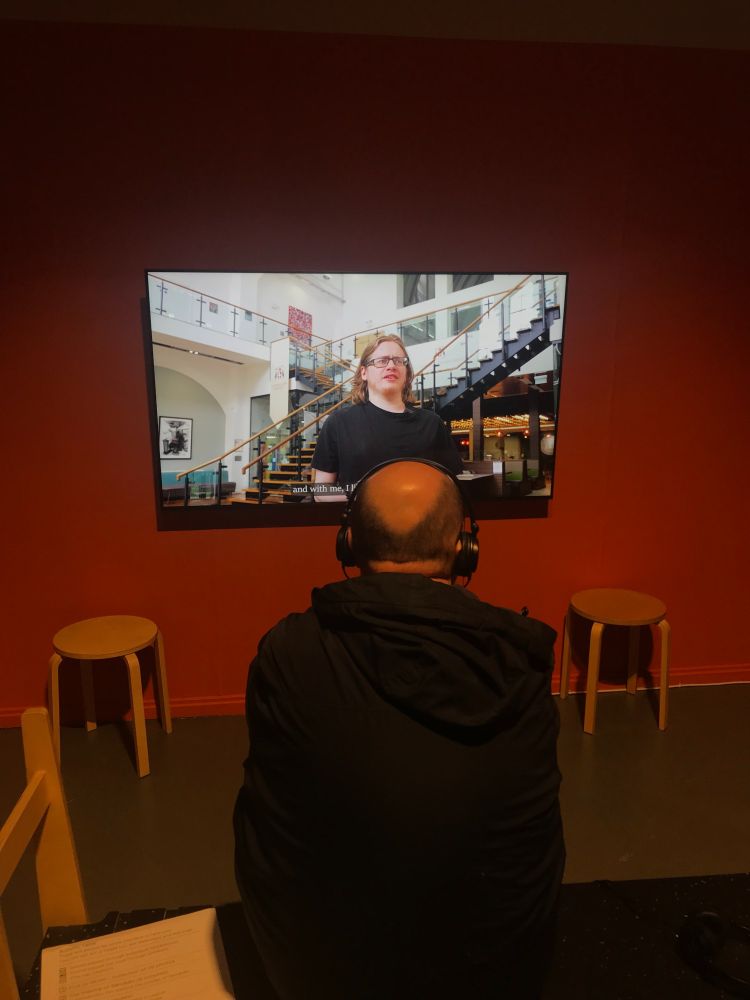 A person wearing headphones sits in front of a screen displaying a video interview. A captioned image of a speaker appears on the screen, with a wooden stool on either side of the setup. The background wall is painted orange, creating a warm atmosphere.