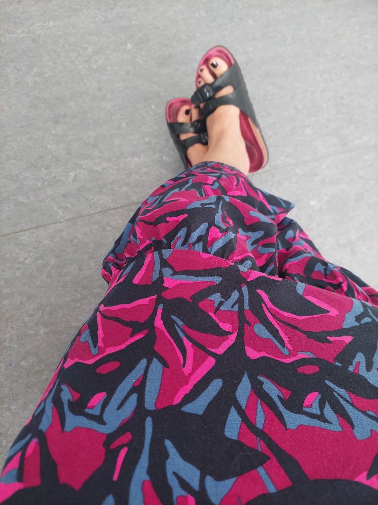 Pink, blau und schwarz gemustertes langes Kleid, aus dem unten Füße in schwarzen Birkenstocks herausschauen.