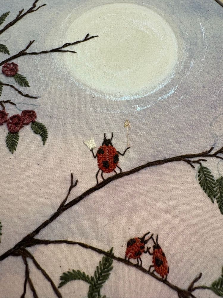 Close up of a WIP, showing little ladybugs having a serenade under the moonlight, surrounded by foliage. One ladybug has a wand and music, and the other two are dancing on the branch below. The ladybugs are made with a basketweave stitch, the leaves with a feather stitch, and the berries with a buttonhole stitch. The moon shimmers with iridescent, glittery watercolor accents. 