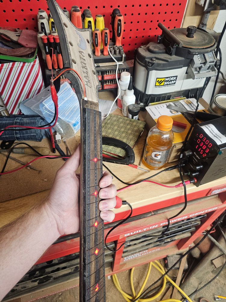 Guitar neck with a light up fretboard hooked to a voltage supply for testing in front of a messy workbench.
