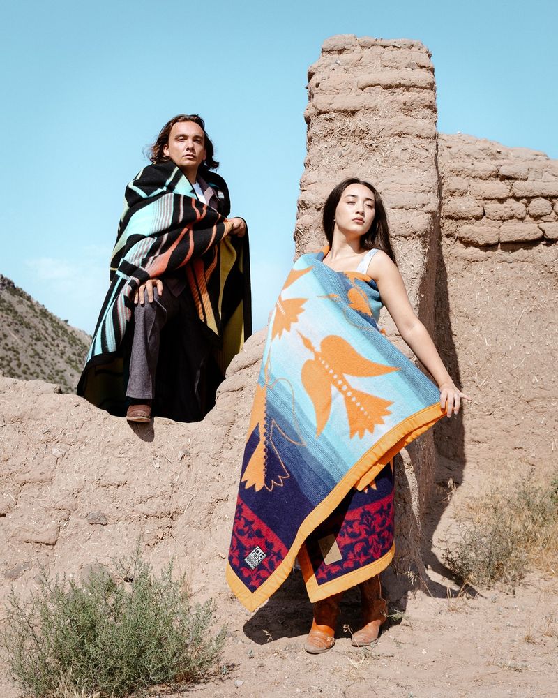 A Native woman models the Legacy Blanket while leading against an adobe structure while a Native man models the Reflection Blanket. 