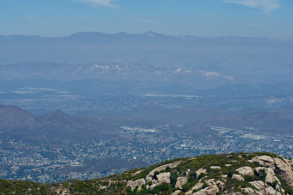 View from Sandstone Peak in the Santa Monicas, southern CA. In the foreground rises a rocky green-beige hillock, but the bulk of the frame is a receding series of mountain ridges, interspersed with the distant, dense geometry of valley settlement. The predominant atmospheric color is a bright, hazy blue.