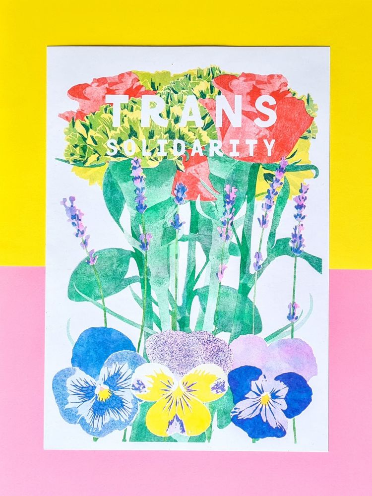 A riso print laying flat on pink and yellow paper. The print features some flowers that symbolise queer identities, pansies at the bottom, lavander in the middle and carnations and roses at the top. The top flowers are broken by white text that reads TRANS SOLIDARITY. The print background is also white. 