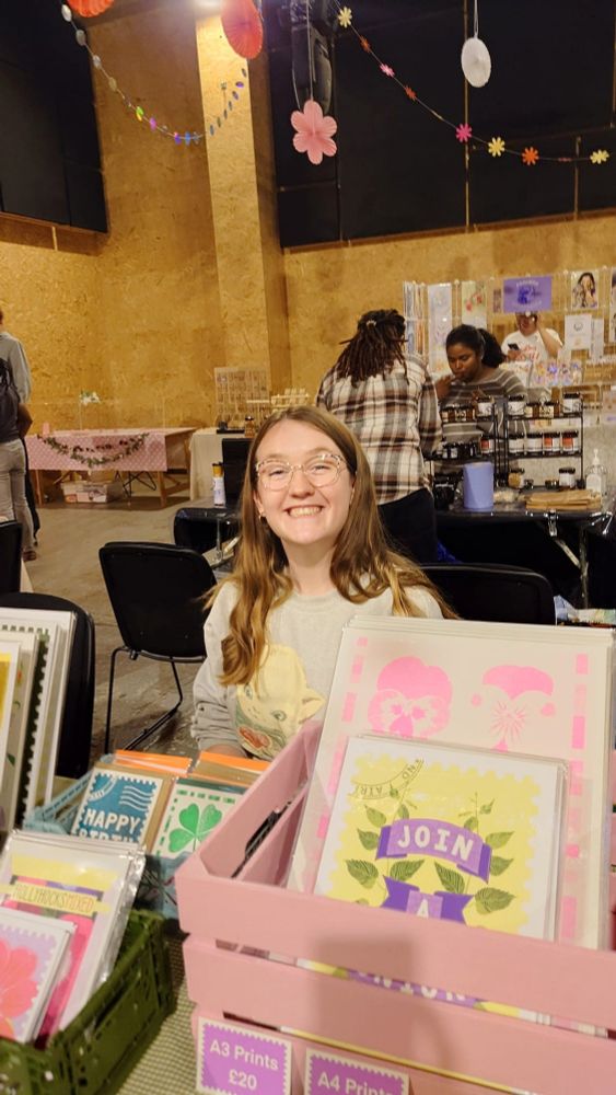 A smiling woman with long light brown hair and clear-framed glasses sits at a market stall filled with colourful illustrated prints and greeting cards. She's wearing a light grey sweatshirt featuring a large graphic of a cat. In front of her are neatly displayed prints, including bright floral and postage stamp inspired designs in pastel colours. Behind her, other vendors and visitors are visible in a warmly lit indoor venue with wooden walls and playful paper decorations hanging from the ceiling. The atmosphere is cheerful and creative.
