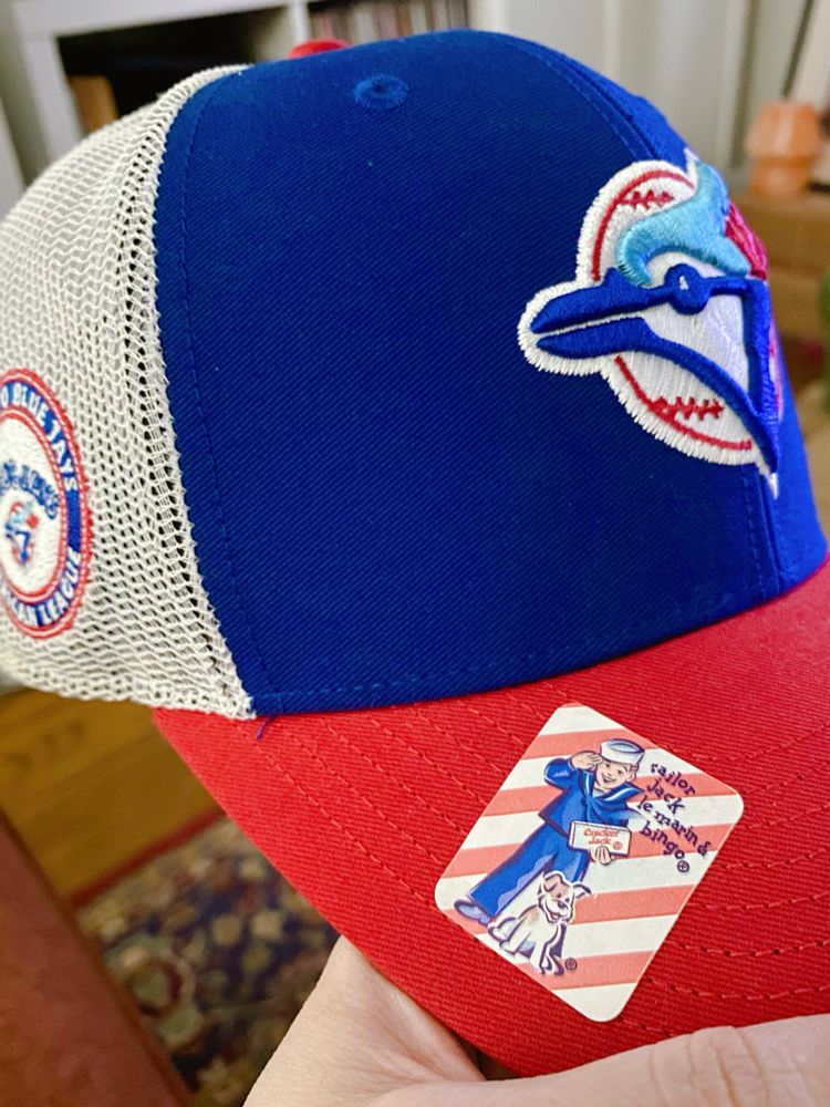Close up photo of a royal blue Blue Jay’s hat with a red brim, decorated with a red and white striped Cracker Jack sticker with a sailor on it