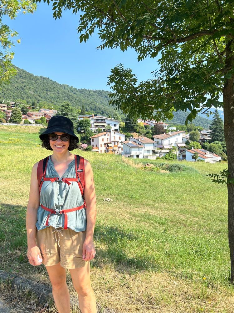 Picture of me, smiling and wearing sunglasses, standing in front of a grassy field with a cluster of red-roofed white houses in the middle ground, and tree-covered hills behind them. I'm in Ranica, a small town in northern Italy.