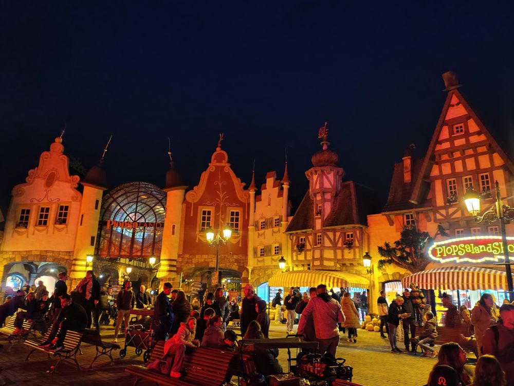 Eingangsbereich des Plopsalands im Dunklen mit in orangenen Licht beleuchteten Buden und Geschäften, davor viele Menschen 